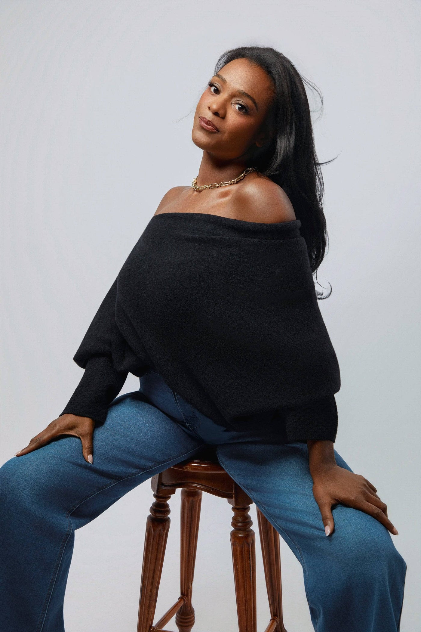 Woman wearing a black off-shoulder top and blue jeans sitting on a wooden stool against a plain background