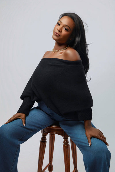 Woman wearing a black off-shoulder top and blue jeans sitting on a wooden stool against a plain background