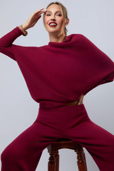 Woman wearing a burgundy outfit sitting on a wooden stool against a plain background