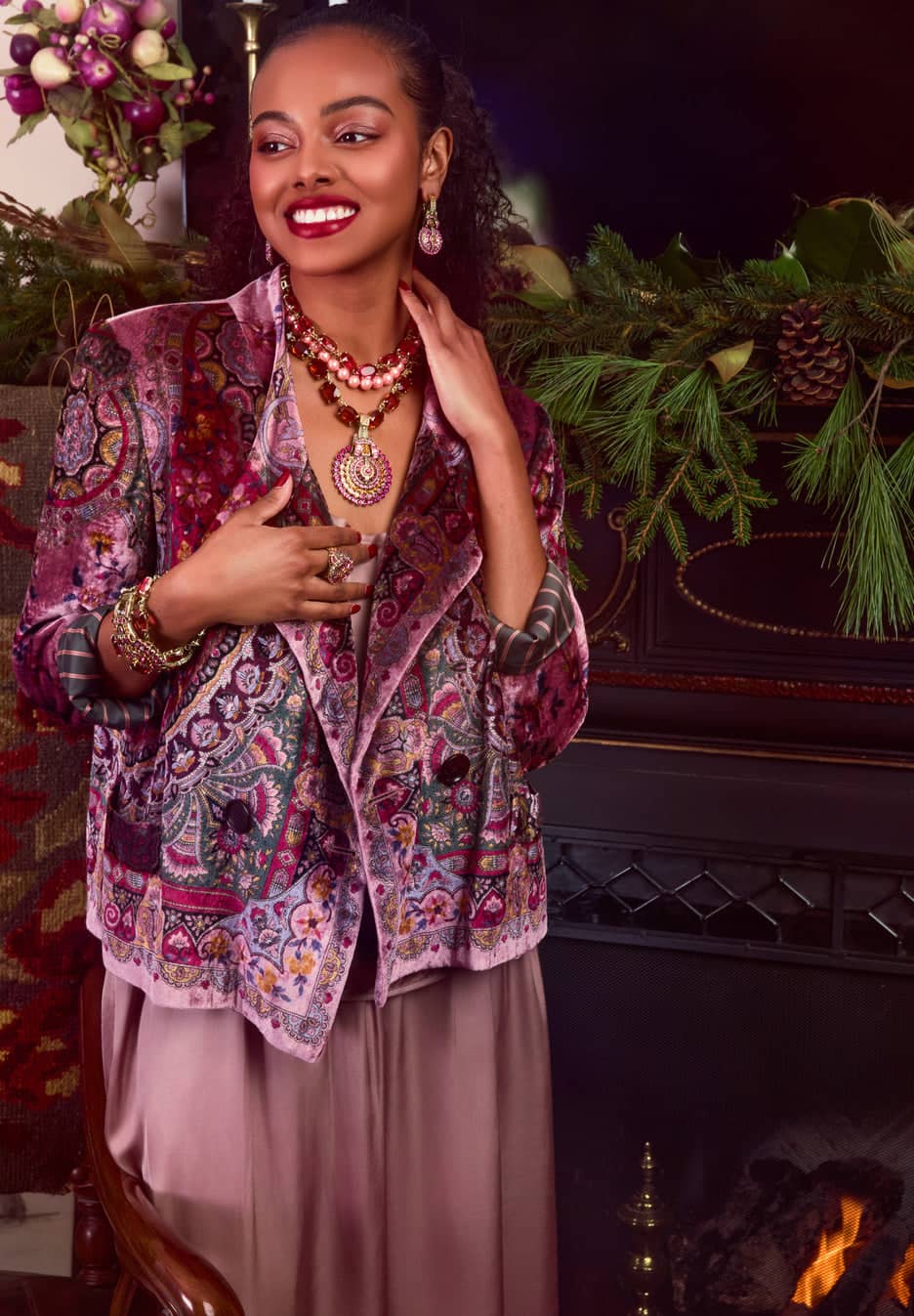 Woman wearing a patterned jacket in a festive indoor setting with greenery and a fire.