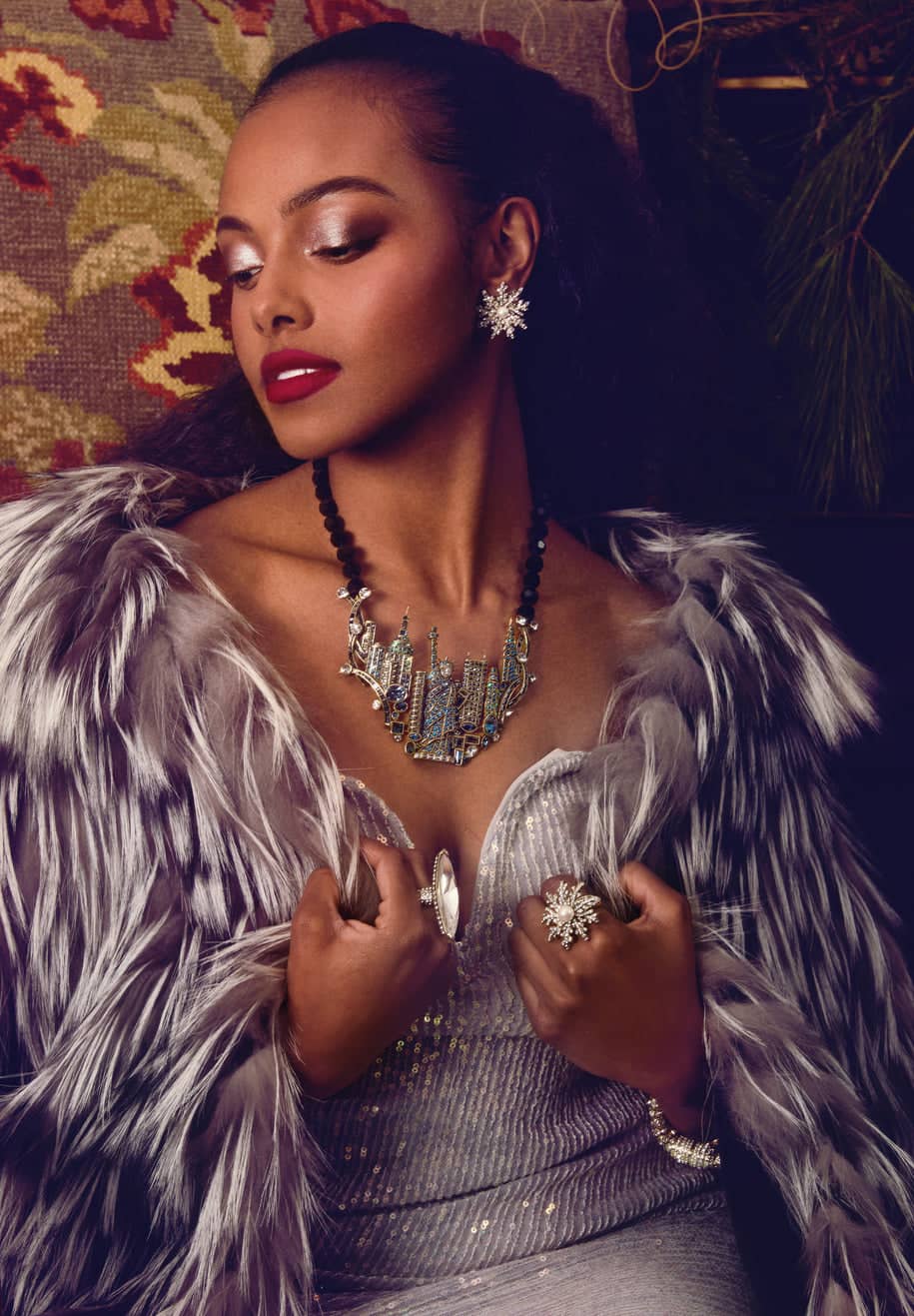 Woman in a glamorous outfit with a feathered garment and jewelry, against a patterned wall.