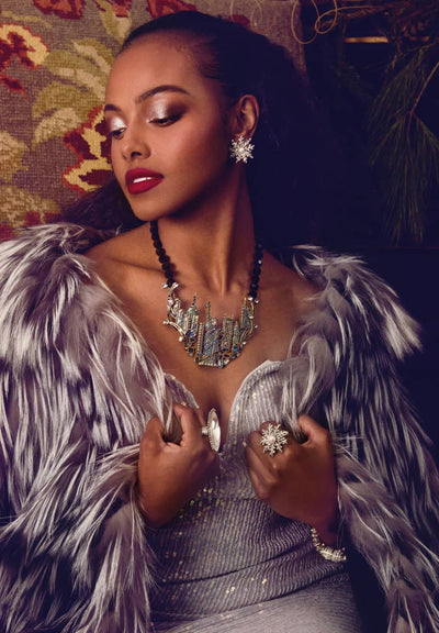 Woman in a glamorous outfit with a feathered garment and jewelry, against a patterned wall.