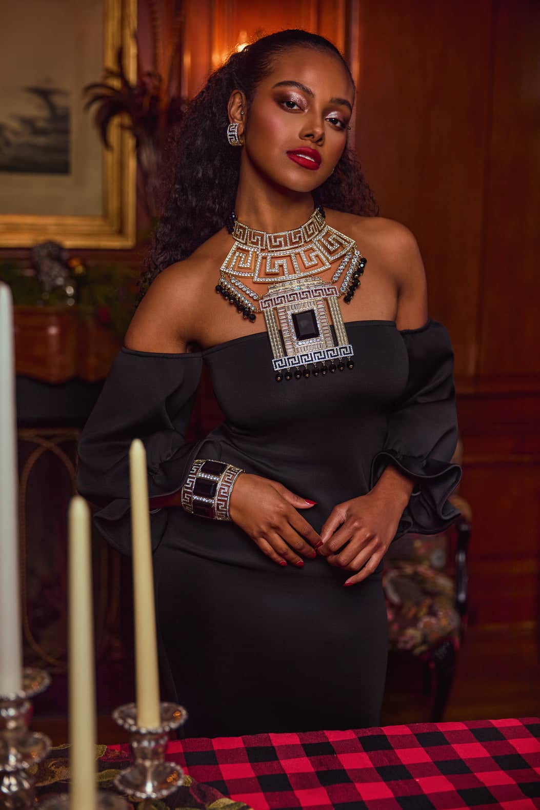 Woman in a black off-shoulder dress with intricate jewelry in an elegant indoor setting.