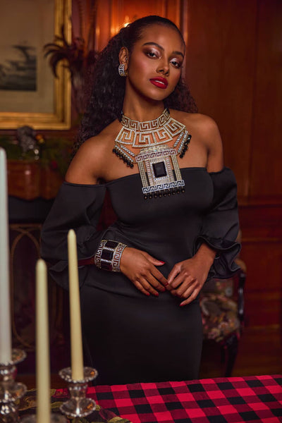 Woman in a black off-shoulder dress with intricate jewelry in an elegant indoor setting.