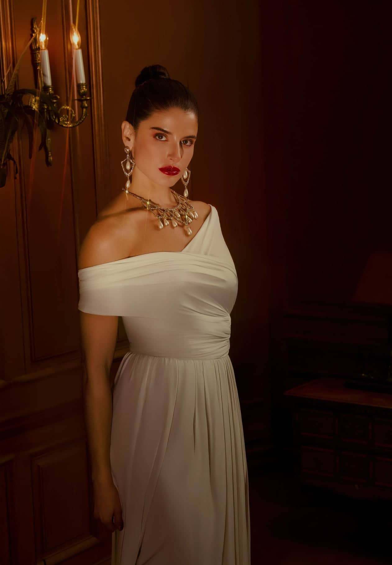 Woman in a white one-shoulder dress with gold jewelry against a warm-toned wall.