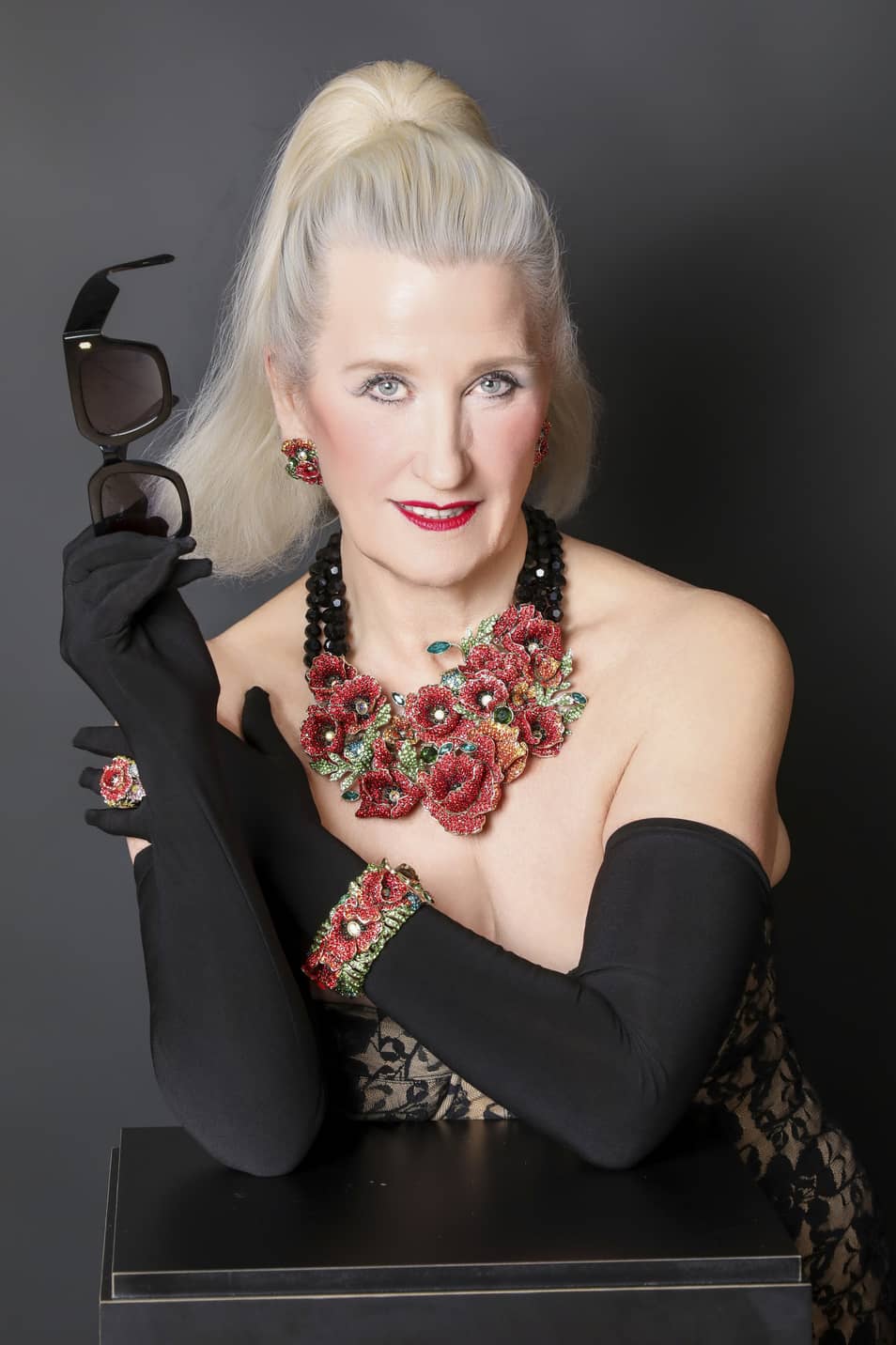 Woman with floral jewelry and black gloves holding sunglasses against a dark background