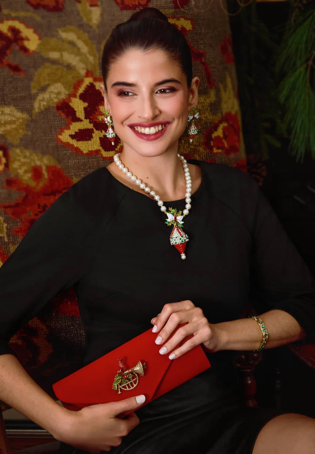 Woman holding a red clutch with jewelry, sitting against a patterned wall.
