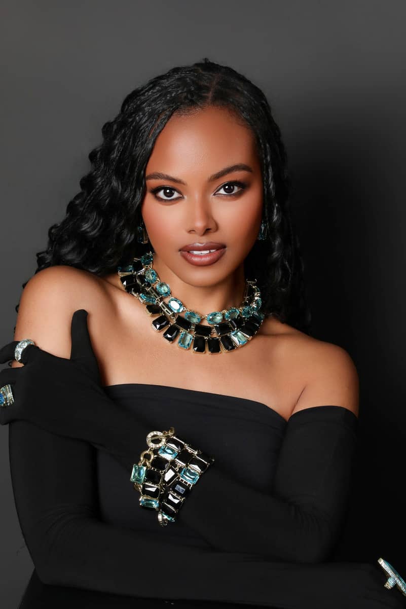 Woman wearing black off-shoulder top and long black gloves with jewelry against a dark background