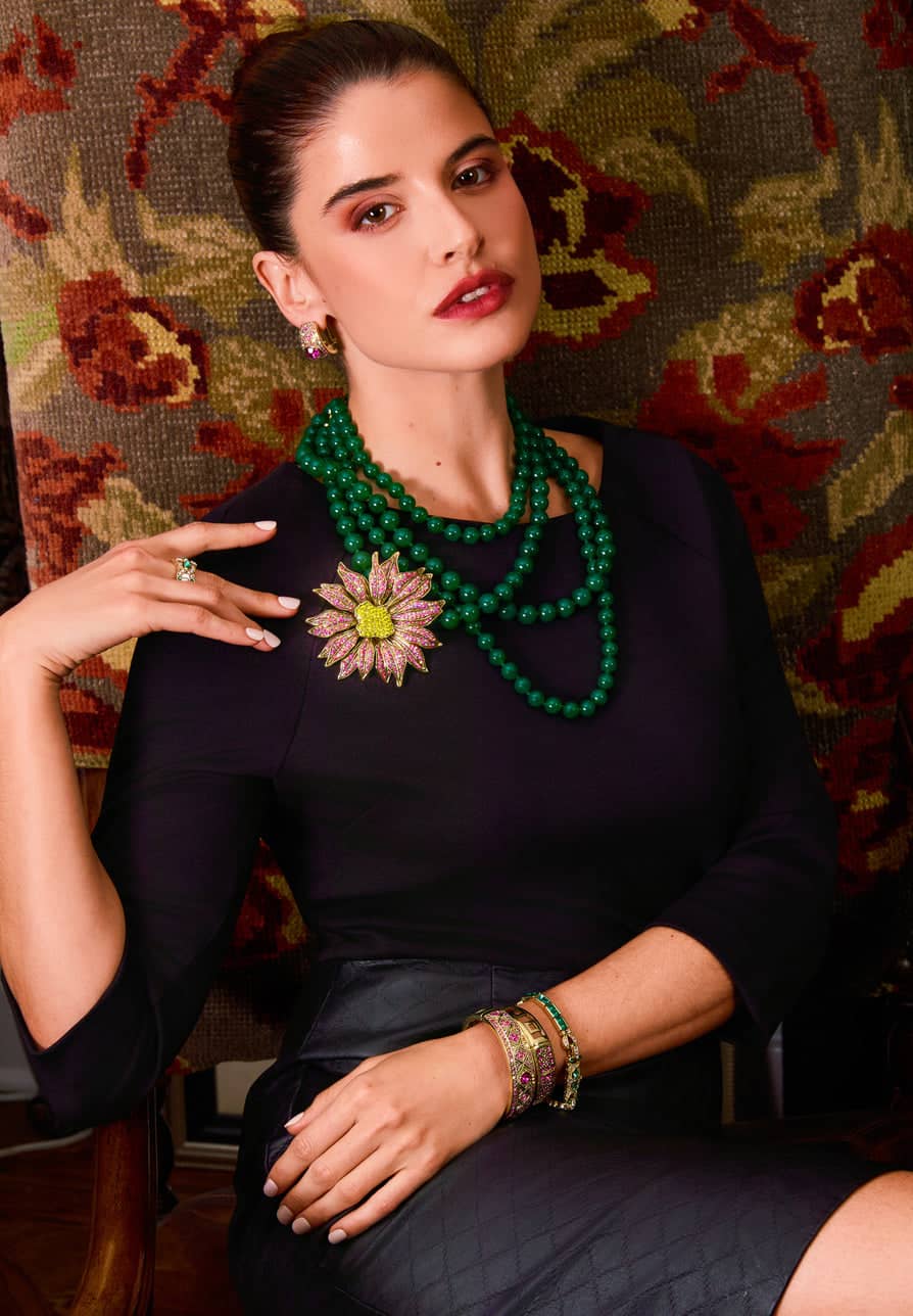 Woman wearing a green beaded necklace and floral brooch against a patterned wall.