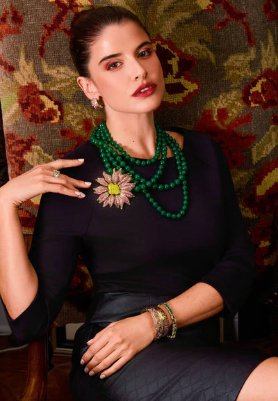 Woman wearing a green beaded necklace and floral brooch against a patterned wall.