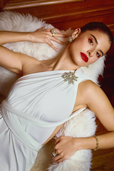Woman in a white dress with feathered details, wearing red lipstick and gold jewelry, indoors.