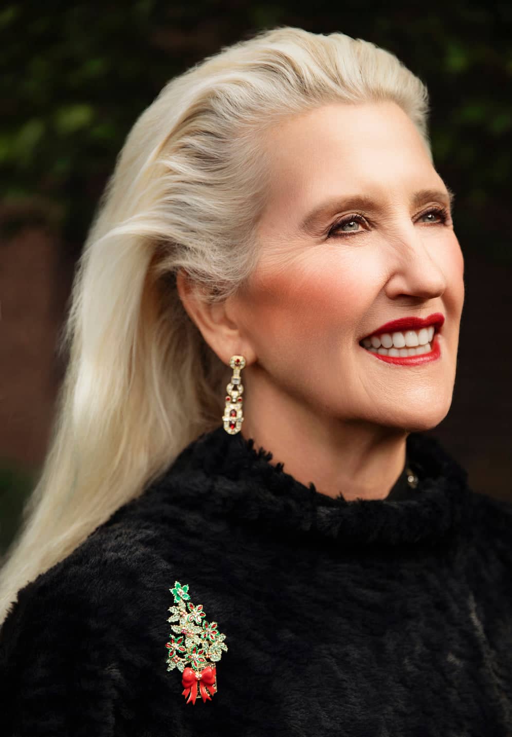 Woman with styled blonde hair wearing a black outfit with a decorative brooch, against a blurred natural background.