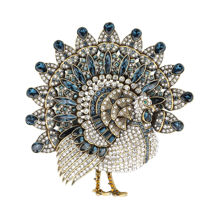 An ornate peacock brooch featuring gold-toned metal, pavé-set clear crystals, and deep blue marquise and round stones, with a small green eye accent, shown on a plain white background.