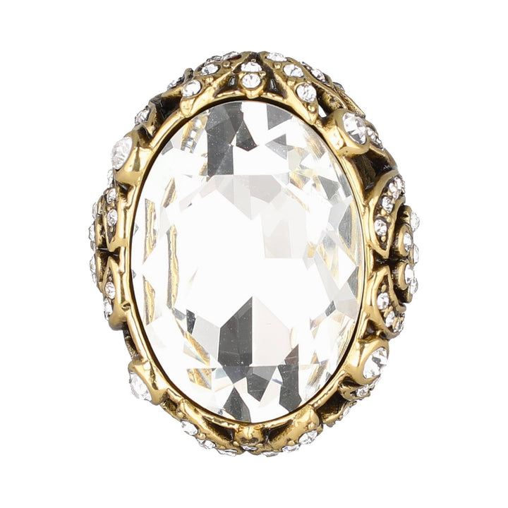 An oval-shaped gold ring featuring a large, clear, faceted crystal gemstone in the center, surrounded by a border of small clear crystals on a plain white background.